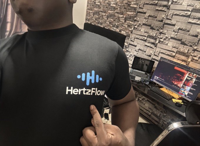 A man taking a mirror selfie pointing at his black t-shirt which features the blue HertzFlow logo, with a laptop and monitor visible on a desk against a stone-textured wall in the background.