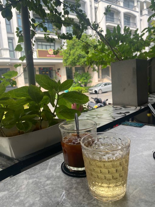Two iced drinks on a cafe table with a view of a street, buildings, and potted greenery.