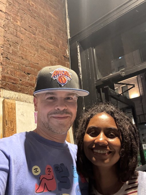 A selfie of a smiling man and woman; the man wears a New York Knicks cap and a light purple t-shirt with abstract faces, standing in front of a brick wall.