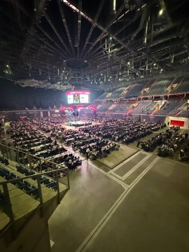 A wide view of a large indoor arena or auditorium with rows of empty seats facing a stage lit with red and white lights.