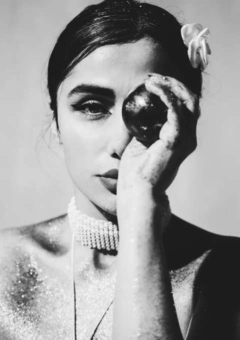 Black and white stylized portrait of a woman with dark hair pulled back, wearing a pearl choker, holding a dark spherical object near her eye, with glitter dusted on her skin.