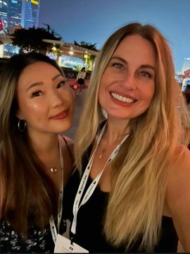 A selfie of two smiling women at an outdoor evening event, with lanyards around their necks and city lights visible in the background.