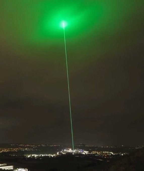 A green laser beam shoots up from a lit structure into the night sky, evoking a "laser eyes" meme aesthetic that suggests an imminent moonshot. The image is used as a dramatic backdrop for the post asking which memecoin is about to experience a massive 10,000x surge.
