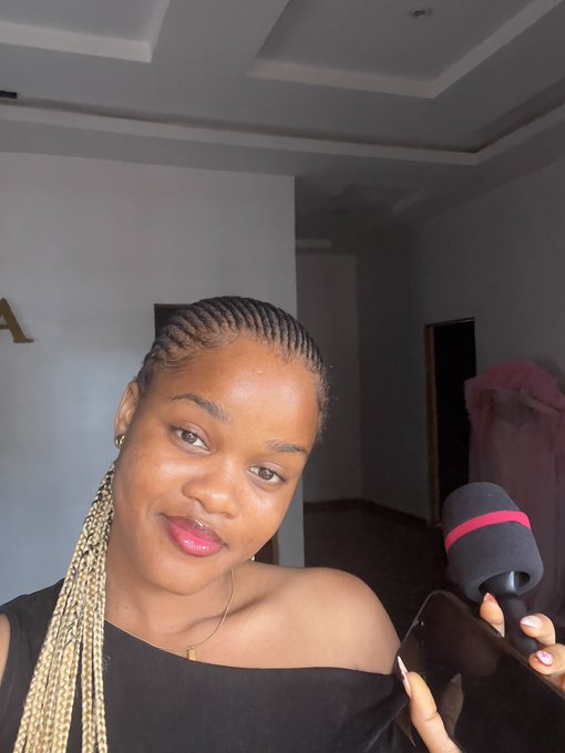 A woman with long blonde braids takes a selfie while holding a phone with a microphone attachment in a room with light-colored walls.