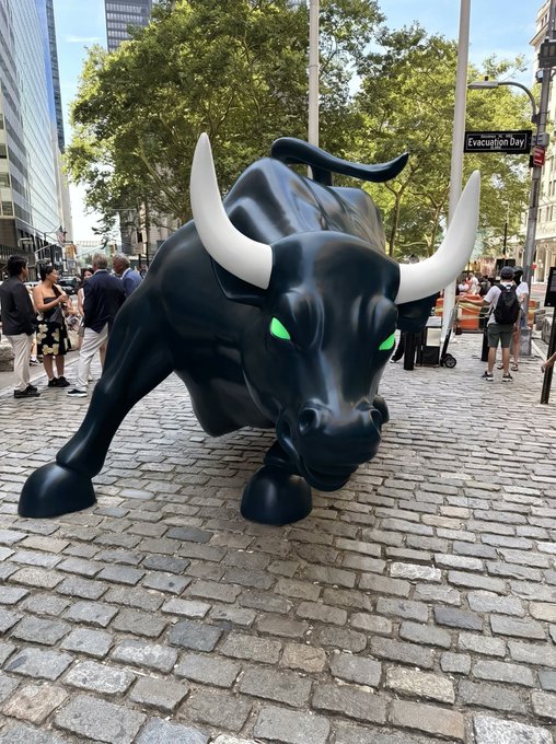 A large black bull statue with white horns and glowing green eyes displayed on a cobblestone street in a city setting.