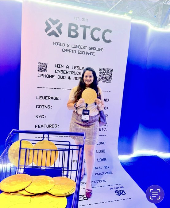 A young woman smiles broadly while holding a gold coin prop in front of a large BTCC promotional backdrop at what appears to be the Token2049 conference. The signage highlights BTCC as "World's Longest Serving Crypto Exchange" (Est. 2011) and promotes a giveaway for "a Tesla Cybertruck, iPhone Duo & more," with a shopping cart full of golden Bitcoin coins completing the playful scene.