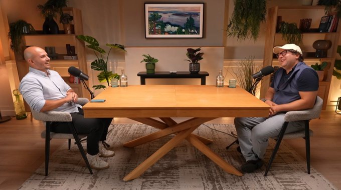 Two men sit across from each other at a wooden table with microphones, recording what looks like a podcast or interview in a warmly lit room decorated with plants and a framed landscape.