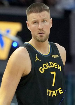 Kristaps Porzingis, wearing a black Golden State Warriors jersey with the number 7, stands on the court with a focused expression. The image directly relates to the breaking news of his indefinite absence due to an unspecified health issue.