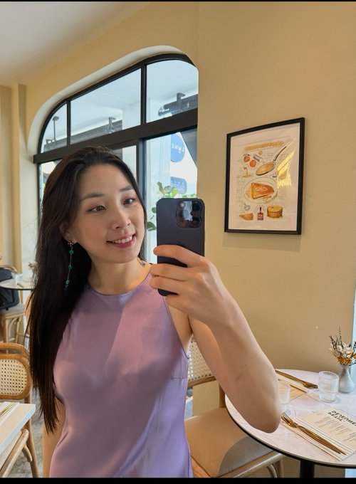 A woman taking a mirror selfie in a cafe, wearing a lavender sleeveless top and dangling earrings.