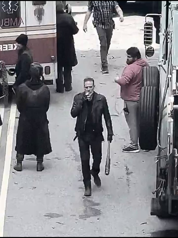 The image shows a man walking down an empty street while smoking a cigarette. A large truck is parked nearby with a trailer attached. There are several other people standing around, including one person wearing a backpack. The scene appears to be related to a story about someone buying real estate for $7050 usingETH chips that were created in 2015 and now selling the property for ETH at $1400.