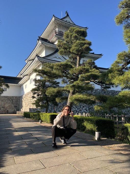 A person squats on a stone path in front of a traditional Japanese castle with white walls, dark roofs, and pine trees against a clear blue sky.