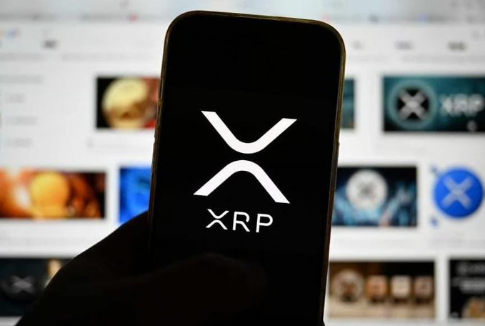 A hand holding a smartphone displaying the XRP cryptocurrency logo, with a blurred screen in the background showing various cryptocurrency listings including XRP.