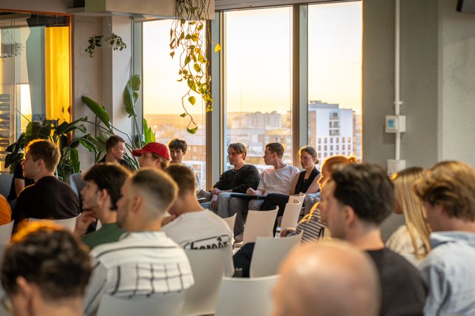 A group of people seated in white chairs listening to a presentation in a bright room with large windows overlooking a city skyline, with green plants hanging from the ceiling.