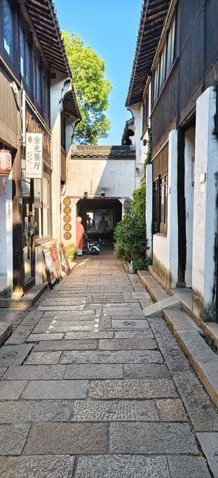 A narrow stone-paved alleyway is framed by traditional Chinese buildings with white walls and dark tiled roofs, featuring hanging signs with Chinese characters such as "朱家厅" and leading to a moon gate.