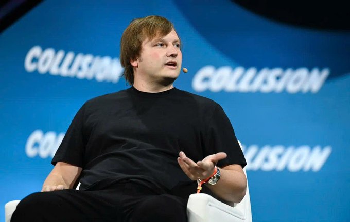 A man in a black t-shirt speaking on stage at a "COLLISION" conference, gesturing with his hand while wearing a wristband and ear-mounted microphone.
