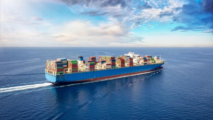 A large blue container ship loaded with multi-colored shipping containers sails across the open ocean under a partly cloudy sky.