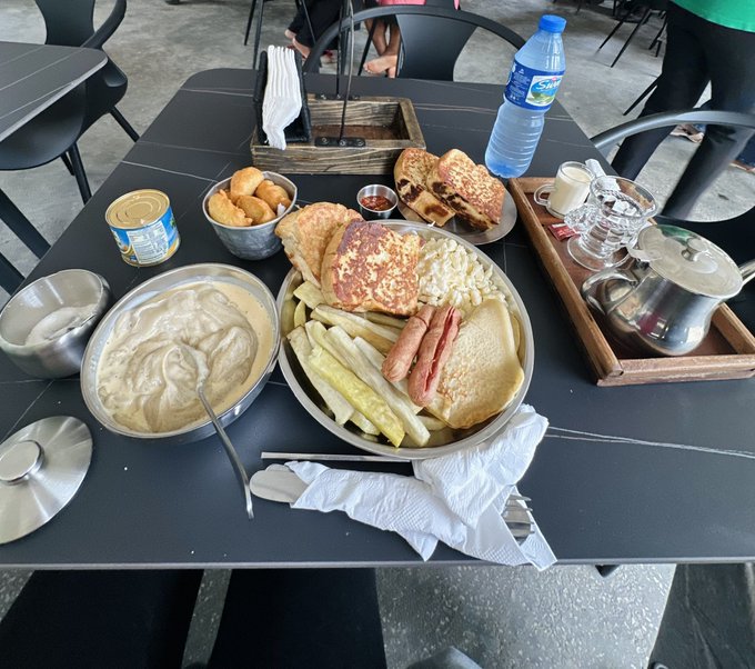 A table set for a hearty breakfast features a large platter piled with French toast, pancakes, scrambled eggs, sausages, bacon, fries, and pickles, accompanied by a bowl of oatmeal or grits, muffins, a water bottle labeled "Nestle Pure Life," and a wooden tray holding a teapot and small glasses. The spread directly matches the post's caption, "breakfast is ready… let's eat," capturing the moment before diving into a generous morning feast.
