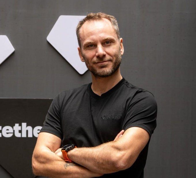 A man in a black t-shirt with the Tether logo stands with his arms crossed in front of a wall with Tether branding, likely representing the company's reported $1.5 billion Q2 net operating profit.