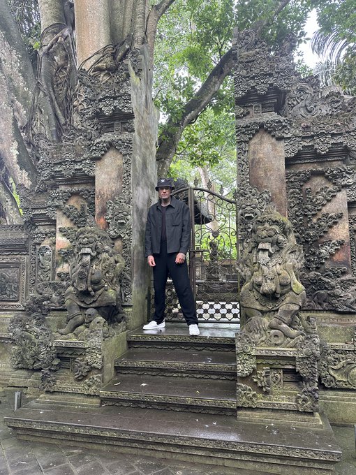 A person in a black outfit and hat standing between two intricately carved stone statues at what appears to be a Balinese temple entrance.