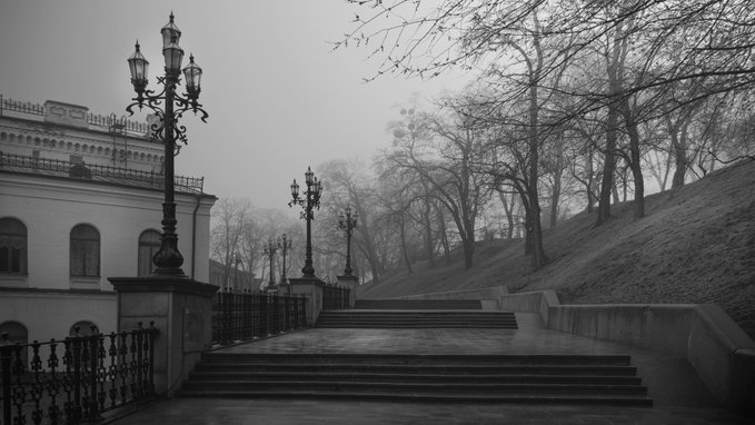 A black-and-white photograph shows an empty stone staircase flanked by ornate lampposts and bare winter trees, with a classical building visible on the left. The monochromatic tones emphasize the misty atmosphere and architectural details, fitting the challenge's "No color. No excuses." theme.