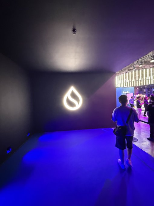 A person walks through a dimly lit, modern hallway illuminated by blue ambient light, approaching a wall with an illuminated logo and a view of a lit-up building outside.