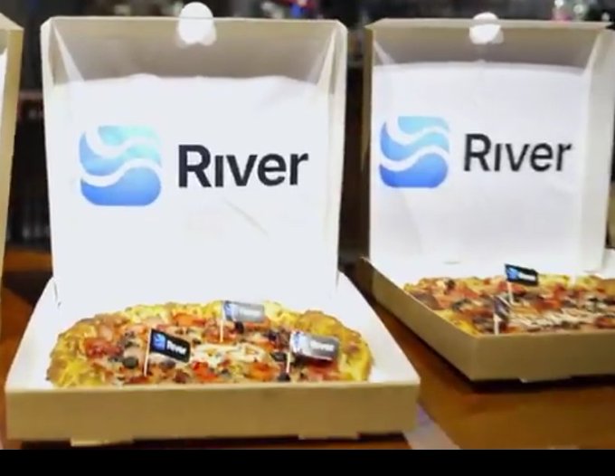 The image features two open pizza boxes placed side by side on a table, each containing a pizza with toppings such as pepperoni, olives, and mushrooms. The word "RIVER" can be seen on one of the pizzas, indicating that they are from the company River. Additionally, there is a small flag attached to one of the pizzas, possibly representing the company's logo or brand.