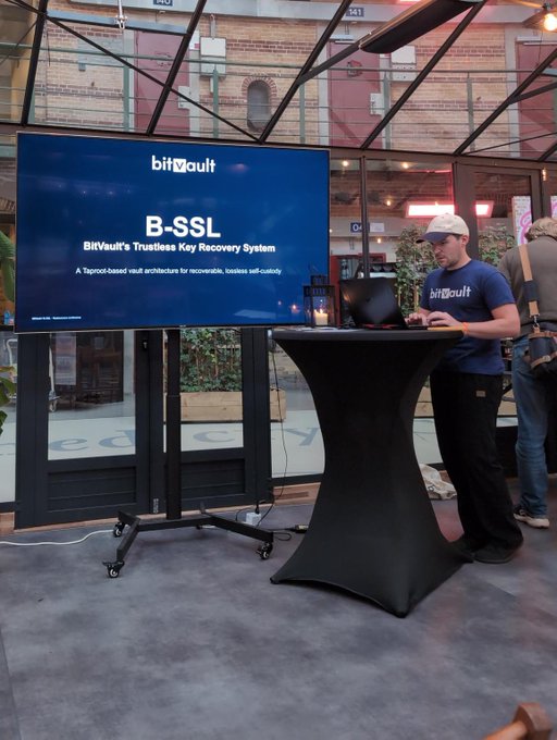 A man in a navy blue 'bitvault' t-shirt and white cap stands at a presentation table with a screen displaying 'bitvault B-SSL: BitVault's Trustless Key Recovery System' at an indoor event venue.