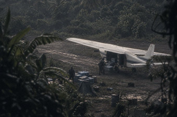 A small white cargo plane sits on a dirt airstrip in a jungle clearing, with armed personnel loading/unloading crates of supplies.