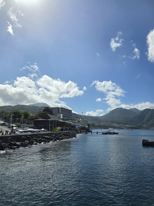 A scenic coastal view features a harbor with buildings, parked cars, and a pier extending into calm blue water, surrounded by lush green mountains under a partly cloudy sky.