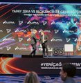 The image shows a stage with two people standing next to each other, possibly giving a speech or presentation. The stage has a large screen behind them displaying text that reads "YAPEY DIKA VE BLOCKOSZÜR BULKÜR BAKSHÇE KARAKAN". There are also several logos visible in the image, including one for "Umarım ilk gün güzel geçmiştir." The stage appears to be set up for an event or gathering.