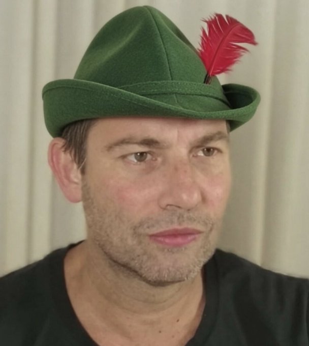 A man wearing a green felt hat adorned with a red feather looks toward the camera against a white curtain background.