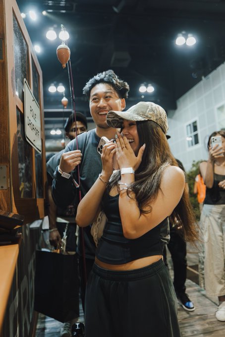 A man and woman laugh joyfully at a phone displaying a Bitcoin (BTC) transaction, presumably the 1 BTC they caught at Lightning Lake, while a sign reading "WE ACCEPT BITCOIN" is visible nearby. The woman covers her mouth in surprise as she holds up her phone to capture the moment, celebrating their crypto catch.