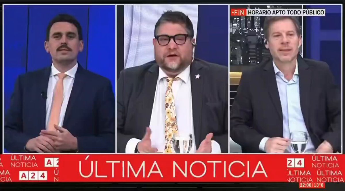 The image shows three men sitting at a table, each wearing a suit and tie. They are engaged in a discussion or presentation, possibly related to National BOLUDO day. The man on the left side of the frame appears to be speaking, while the other two men listen attentively. A TV screen is visible behind them, displaying information about National BOLUDO day.
