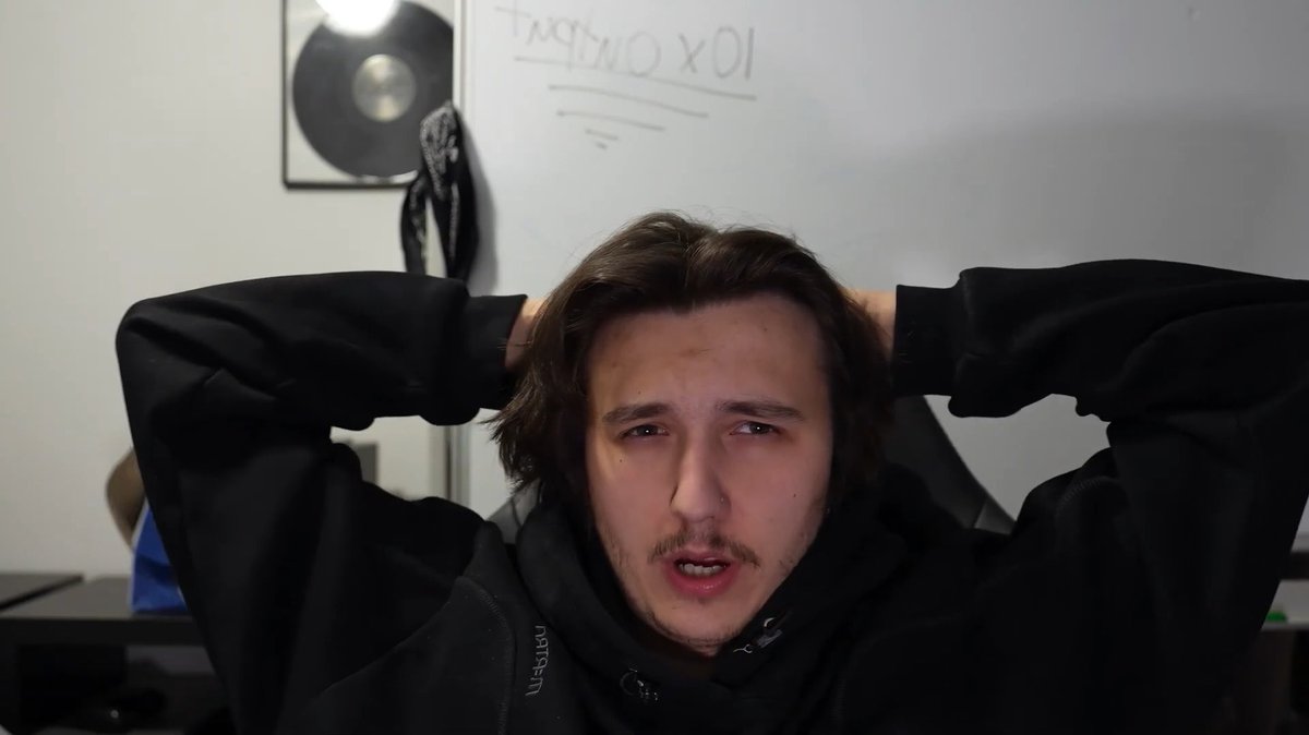 A young man with shoulder-length brown hair and a mustache wearing a black hoodie, sitting with his hands behind his head in front of a whiteboard with '10 x ONLYFANS' written on it and a framed gold record on the wall.
