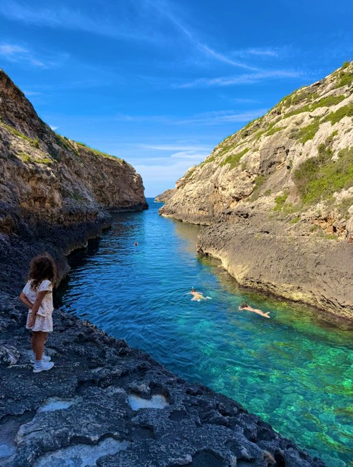 A young girl in a floral dress stands on rugged coastal rocks, gazing out at swimmers floating in a crystal-clear turquoise cove nestled between towering cliffs. The serene, secluded paradise captures the ultimate reward for hard work—a perfect, hard-earned escape into nature's beauty.