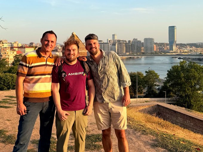 Three men stand together posing for a photo on a hill overlooking a city skyline with a river, with the man in the center wearing a maroon shirt labeled 'AMBIRE'.