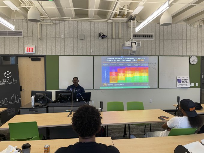 A classroom setting where a Detroit Blockchain Collective event is taking place; an audience member faces a presenter and a large screen displaying a Hive Systems chart titled 'Time it takes a hacker to brute force your password in 2026'.