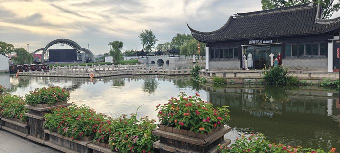 A scenic view of a traditional Chinese garden featuring a pond with stone bridges, a pavilion with curved roof, and vibrant red flowers in the foreground.
