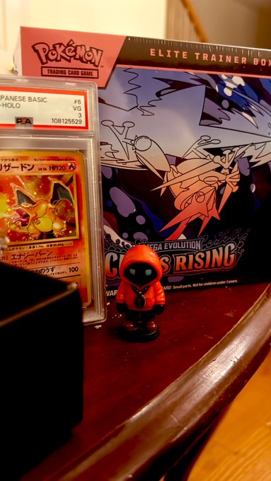 The image shows a Nintendo Gacha toy displayed next to a Pokemon card, both set up on a table or desk. A toy figurine of an elf or wizard wearing a red hood is also present in the scene. The post about Pokémon cards talks about the new "Eliote Rising" board game and how it is collecting and trading cards from various players.