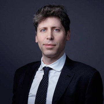 A formal portrait of a man with messy brown hair wearing a dark suit and polka dot tie against a dark background.