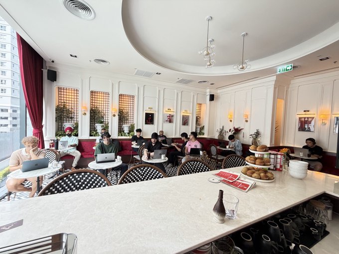 The image shows several people sitting at tables with laptops open in front of them. The room has white walls and ceiling, which gives it an airy feel. There are multiple chairs placed around dining tables, indicating a social setting for work or study. A few potted plants can be seen throughout the space, adding some greenery to the environment. 

The post features a photo of a coffee table with various items on it, including wine glasses and bottles. The image also includes text that reads "Focus on what is in the image and how it relates to the post @Solana_TH".