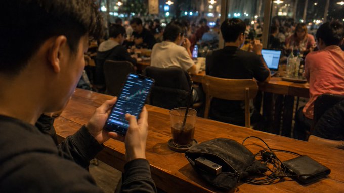 A young man sits at a wooden café table holding a smartphone displaying cryptocurrency price charts, with an iced drink, a black pouch with cables, and another phone resting on the table amid a busy crowd of patrons working on laptops behind him.

The image visually echoes the post's warning about memecoin trading: the solitary trader absorbed in his phone, surrounded by crowds also glued to their screens, mirrors the herd-driven psychology of crypto speculation, while the casual café setting underscores how liquidity (not depth of analysis) is what drives price breakouts in volatile pools.