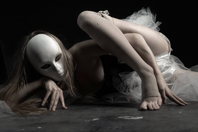 An artistic photo of a person wearing a white mask and tulle skirt, lying on the floor in an elegant pose with white tights.