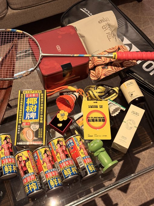 An overhead shot of a glass table displaying a badminton racket, canned coffee with Japanese text, coconut products, and various packaged snacks.