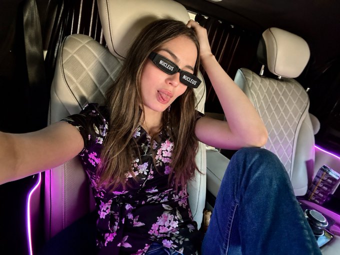 The image shows a woman sitting in the back seat of a luxury car with white quilted leather seats and pink ambient lighting, wearing sunglasses with "NUCLEUS" printed on both lenses and a floral top while taking a selfie and playfully sticking out her tongue. The "NUCLEUS" branding on her eyewear directly ties into the post's mention of @NucleusCodes, visually reinforcing her endorsement of the platform running the campaigns for @sleepagotchi, @CNPYNetwork, and @EthraShip.