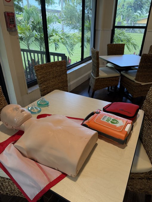 A CPR training dummy lies on a wooden table next to an orange AED defibrillator kit and a face mask, set in a bright room with wicker chairs and large windows looking out to a tropical landscape.