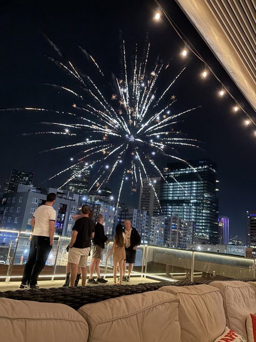 A nighttime rooftop scene where a group of people watch a large fireworks display over a city skyline, with outdoor lounge seating in the foreground.