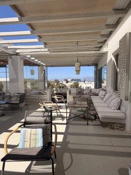 A photograph of a sunny, covered outdoor patio or terrace with white wicker furniture, striped cushions, and large windows overlooking a city landscape.