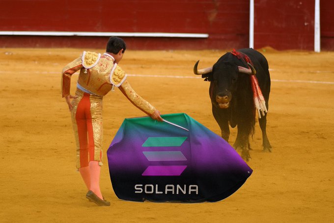 A matador in traditional gold and red attire faces a black bull in a bullring, holding a Solana logo cape and a sword.