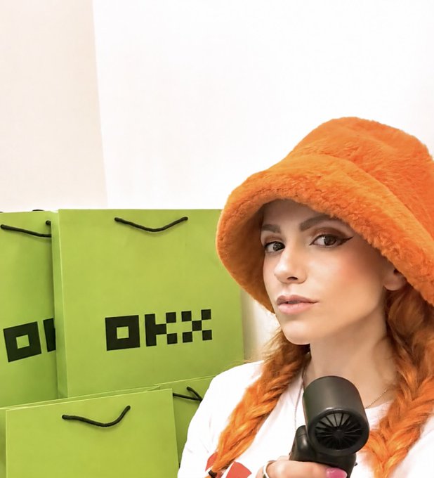 A woman in an orange bucket hat holds a microphone in front of green shopping bags with pixelated designs.